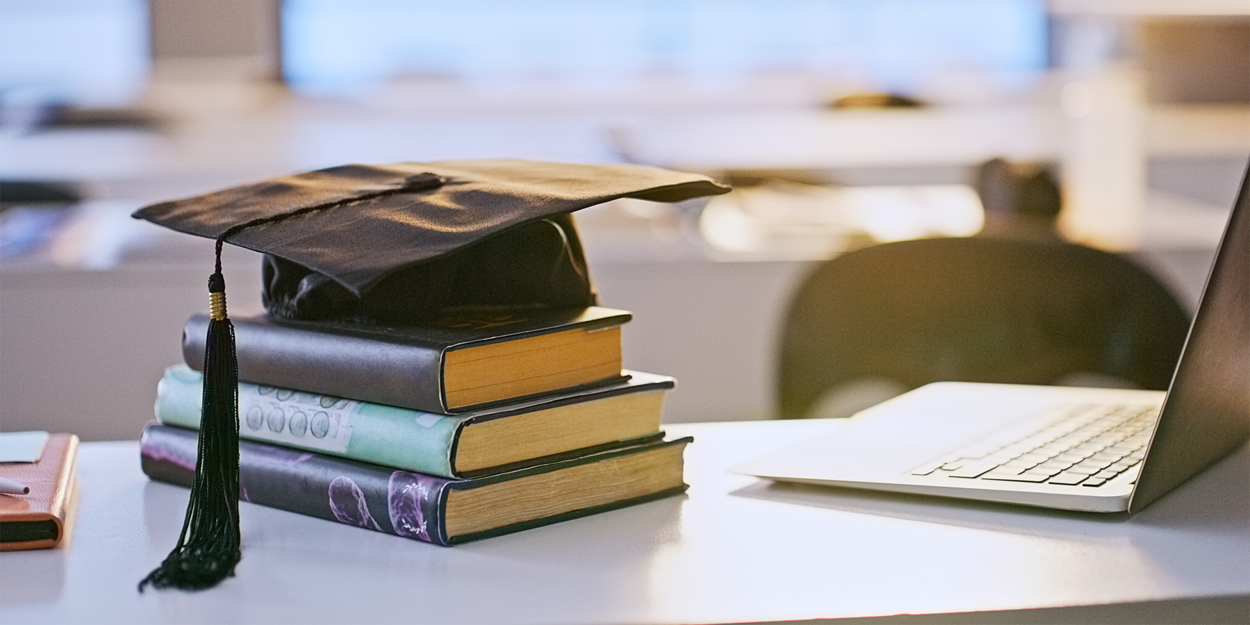 Stack of books with a grad cap on top lying next to an open laptop.