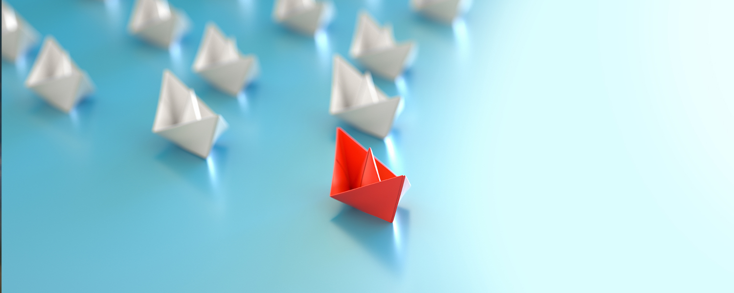 A red paper boat leading several white paper boats. The white paper boats are slightly blurred as they are not in focus.