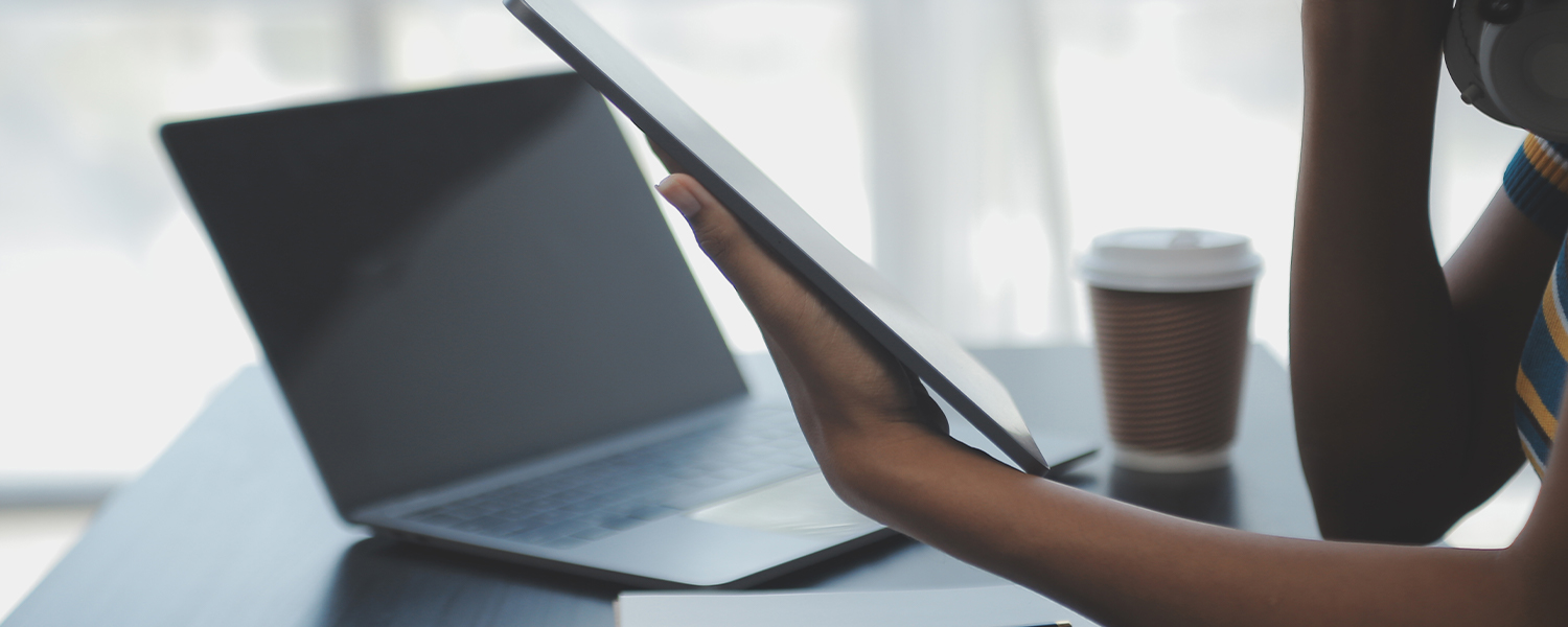 A woman is looking at a tablet with a laptop open in the background.