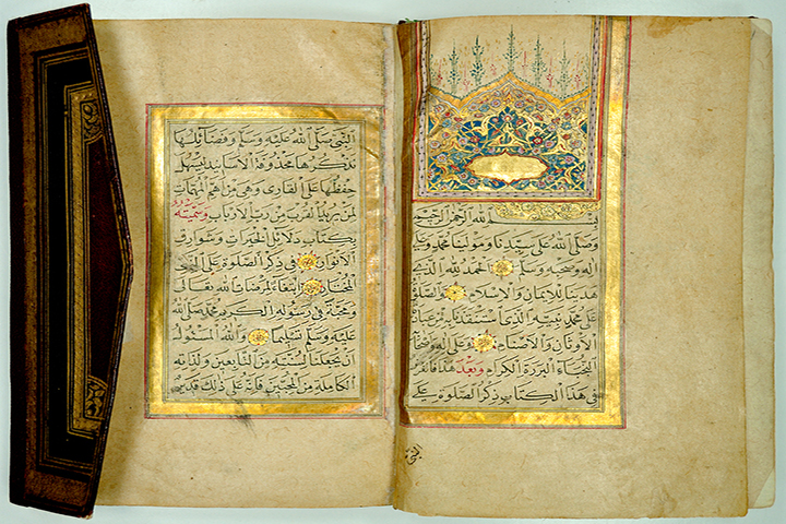An open Islamic prayer book, introduced by a gilded decorative panel with an arch-shaped ornament above. Part of the pointed envelope flap binding is visible.