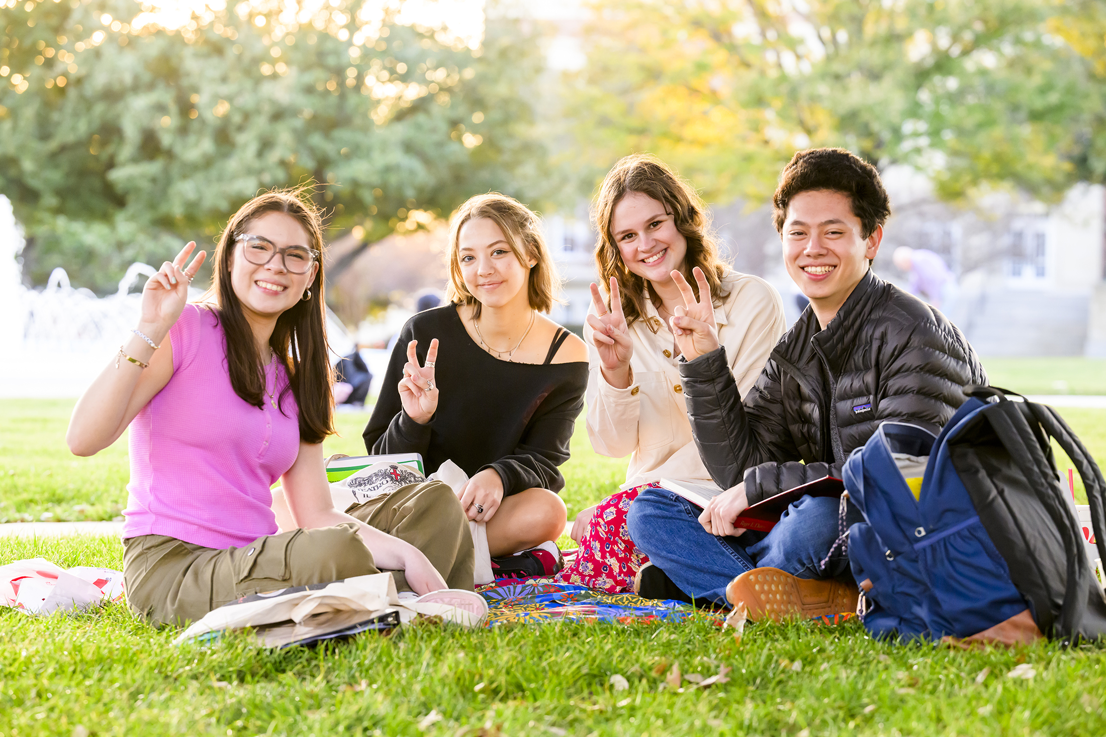 Four students are relaxing on the grass at 色花堂_色花堂 98堂, smiling and holding up their fingers in the University's traditional "pony up" greeting.