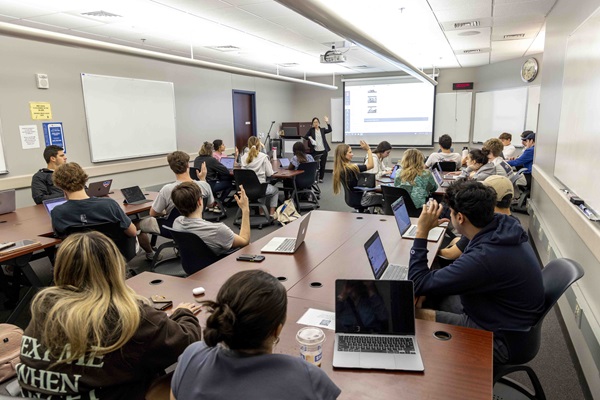 A college classroom with students seated around large, rectangular tables, engaged in a lecture. Many students have laptops open, and some are raising their hands. A professor, standing at the front near a projector screen displaying a slide, is leading the discussion. The room has whiteboards on the walls, a clock, and instructional posters. The setting is well-lit with a professional learning environment.