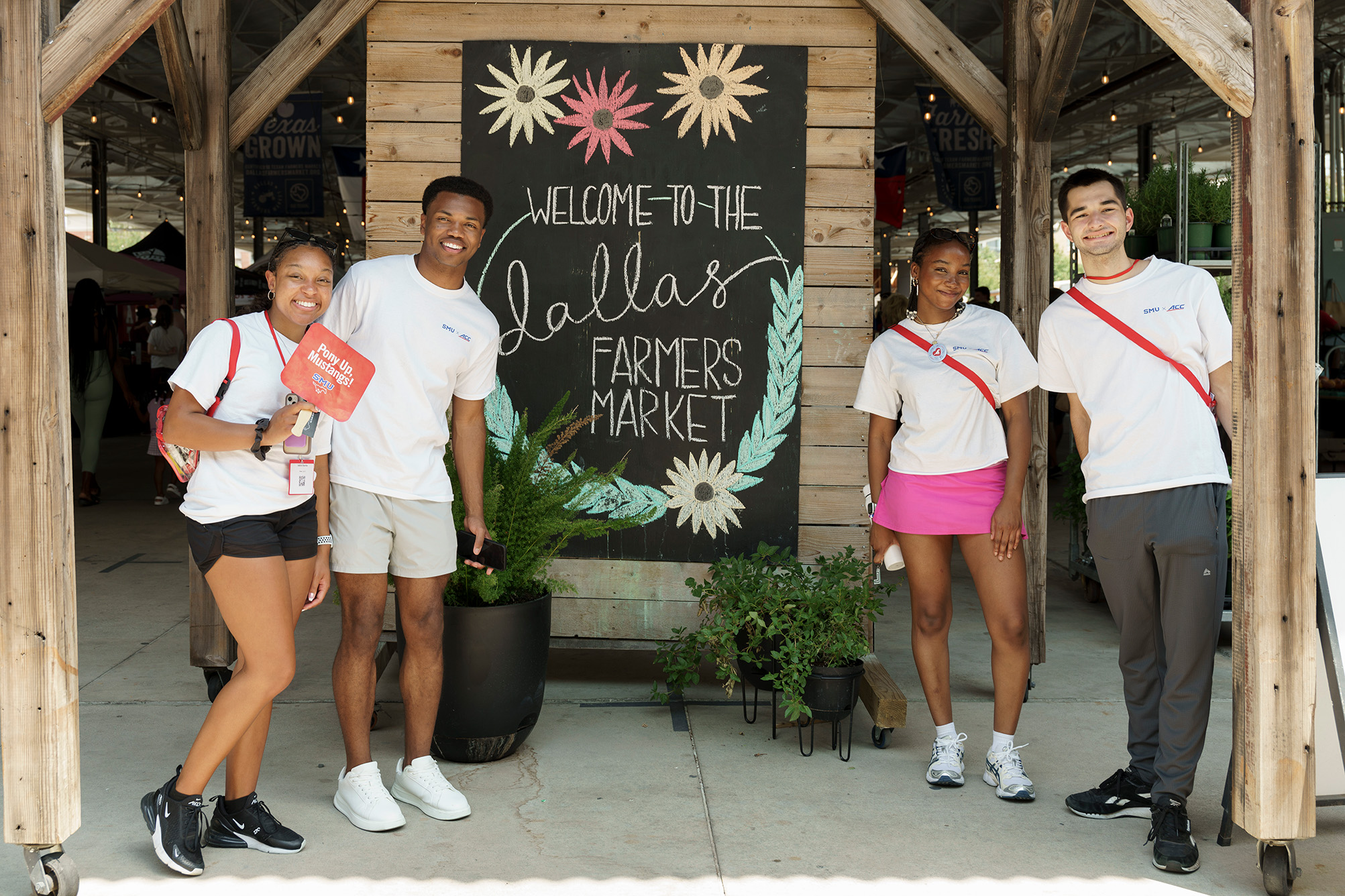four students standing outside in white 色花堂_色花堂 98堂 t shirts in front of dallas farmer's market sign